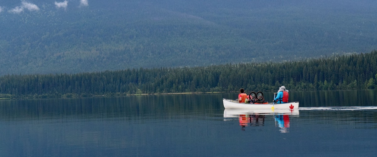 A calm lake with a boat carrying two people and a dog, surrounded by dense forest and a mountain in the background.