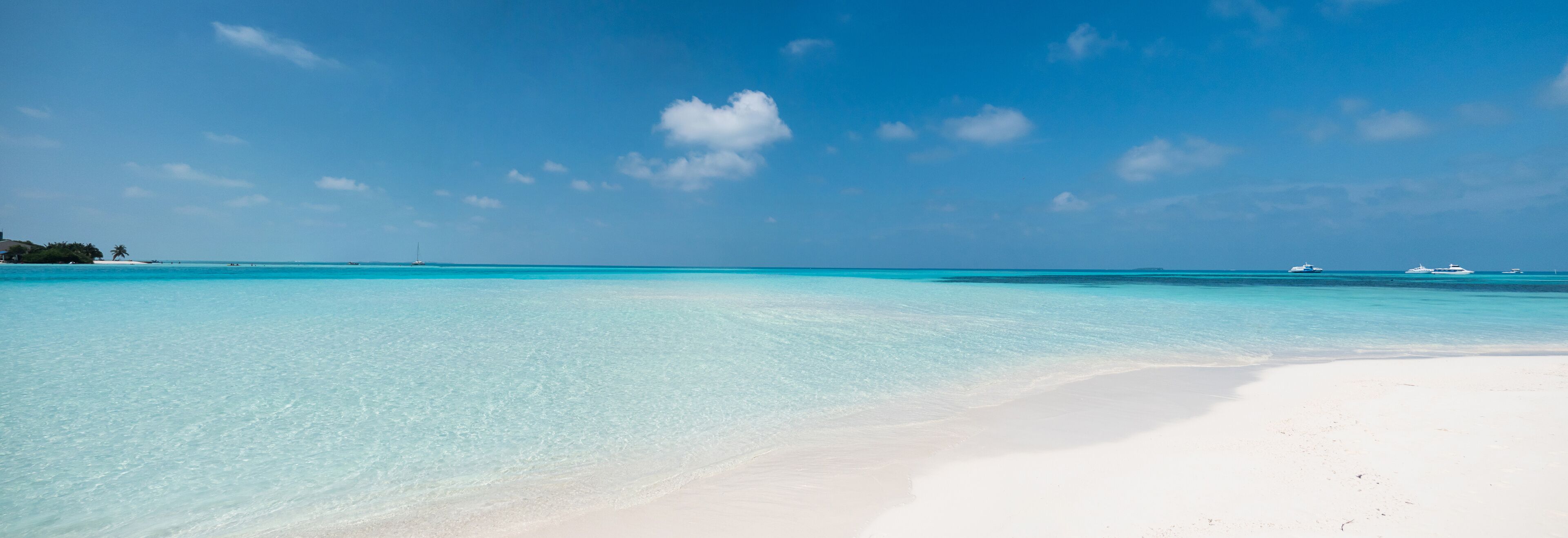 A beach with clear turquoise water, white sand, and a few boats in the distance.