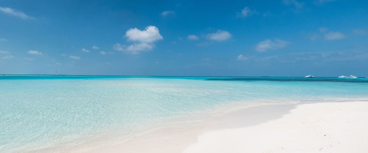 A beach with clear turquoise water, white sand, and a few boats in the distance.