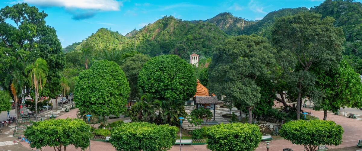 A lush green park with a pagoda in the center, surrounded by dense trees and hills in the background.