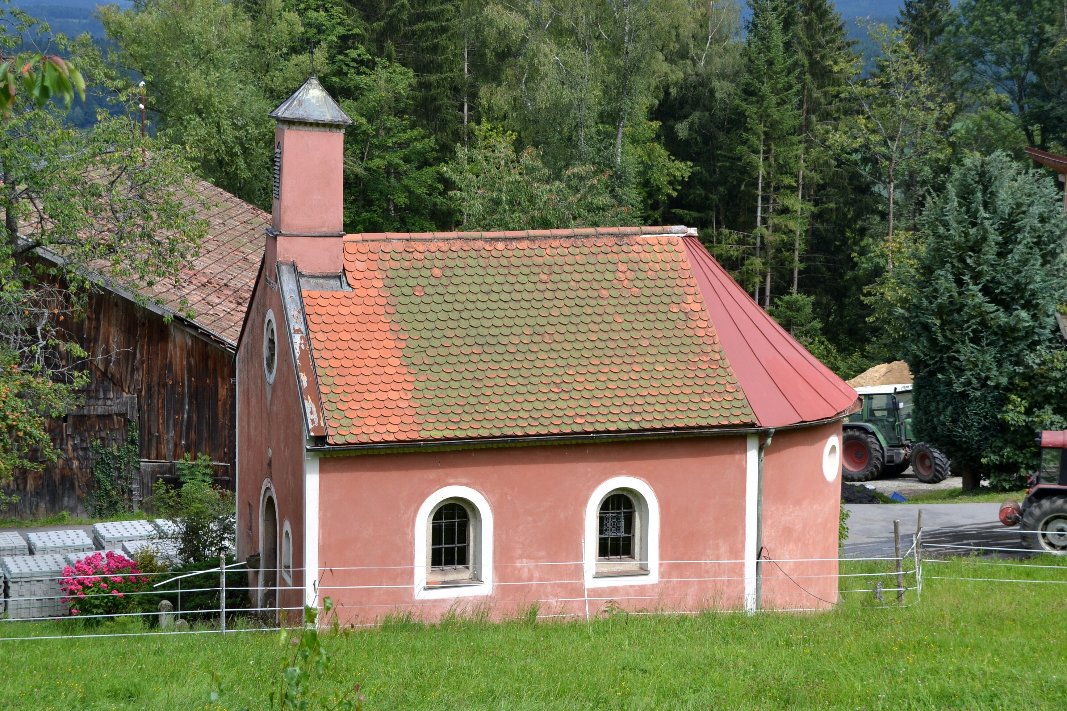 A small red brick building with a green roof, surrounded by lush greenery and a wooden barn.