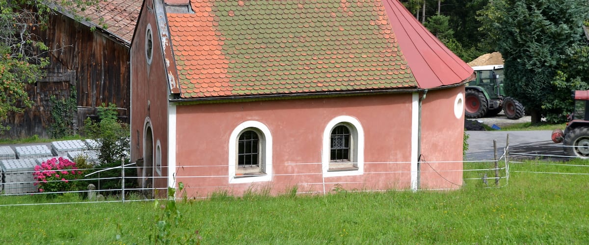 A small red brick building with a green roof, surrounded by lush greenery and a wooden barn.