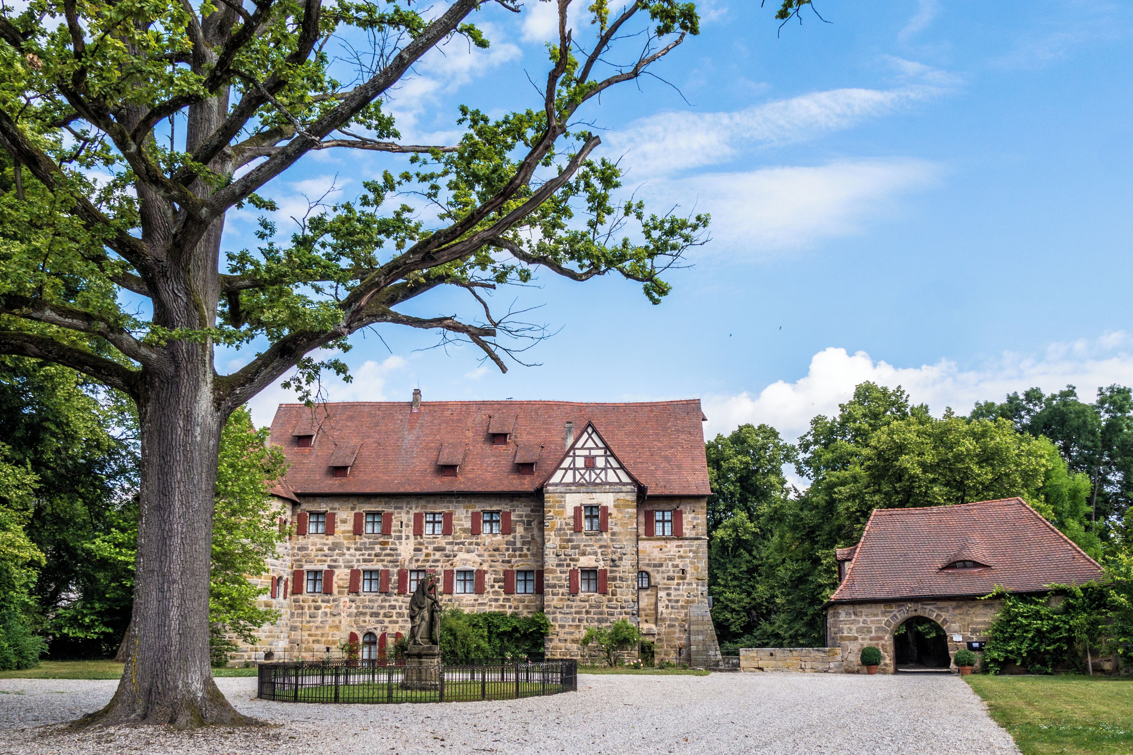 A historic stone building with a red-tiled roof, surrounded by lush greenery and a large tree.