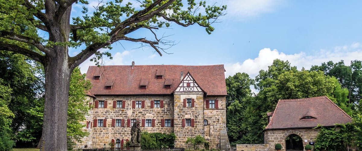A historic stone building with a red-tiled roof, surrounded by lush greenery and a large tree.