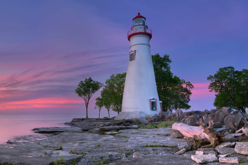 Un phare blanc, coiffé d’une partie rouge, se dresse sur une côte rocheuse au coucher du soleil.