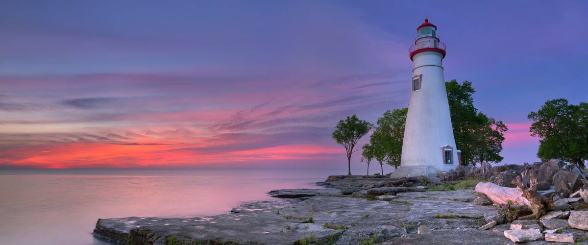 A white lighthouse with a red top on a rocky shoreline at sunset.