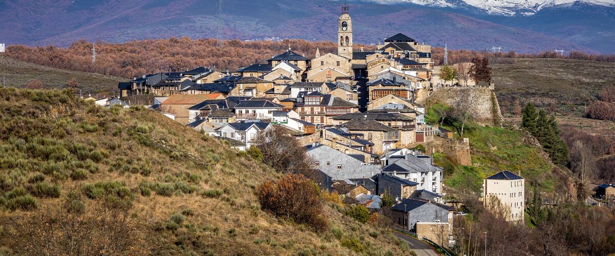 A historic town with a prominent church tower, surrounded by snow-capped mountains and autumn-colored trees.