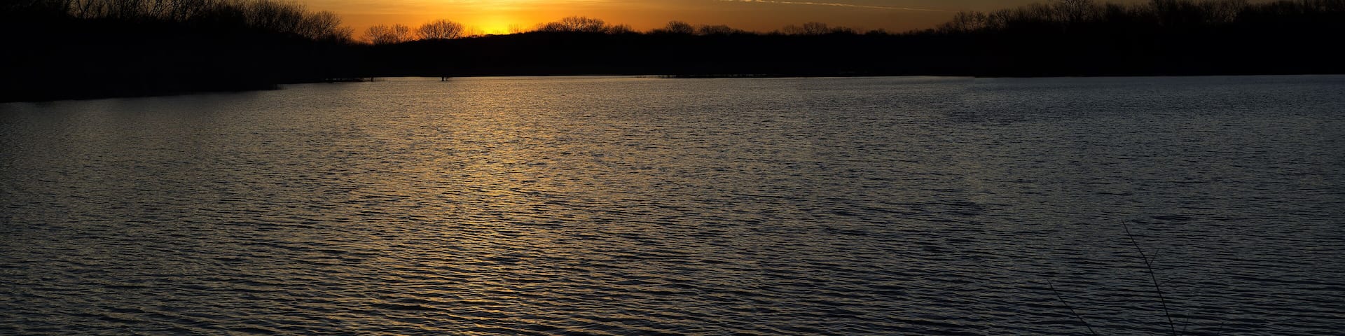 A sunset over a calm lake with silhouettes of trees and a clear sky.