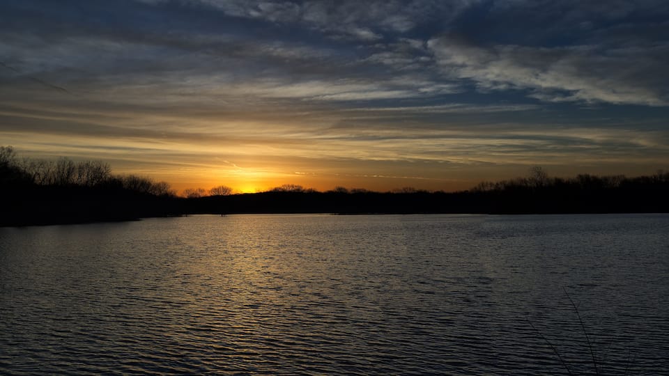 A sunset over a calm lake with silhouettes of trees and a clear sky.