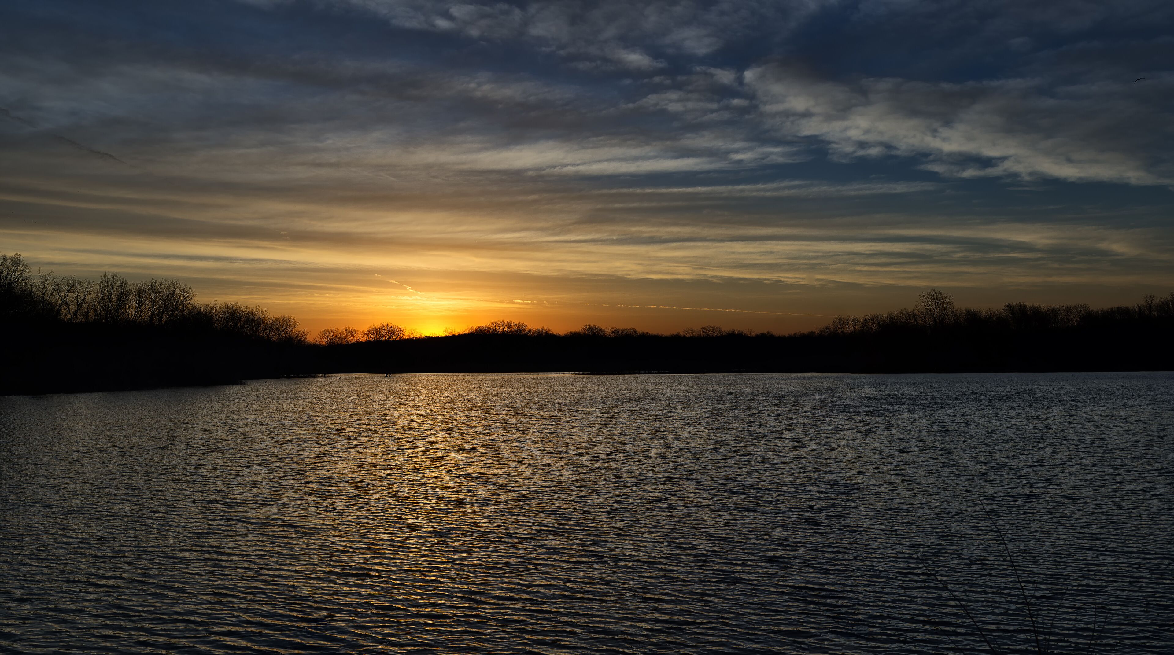 A sunset over a calm lake with silhouettes of trees and a clear sky.