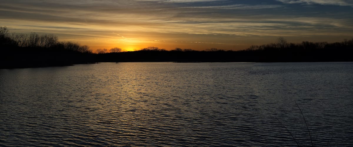 A sunset over a calm lake with silhouettes of trees and a clear sky.