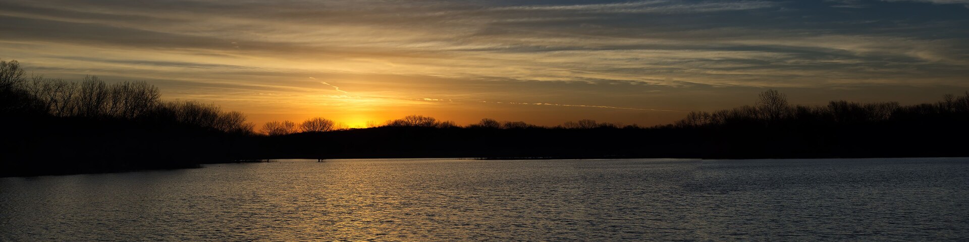 A sunset over a calm lake with silhouettes of trees and a clear sky.