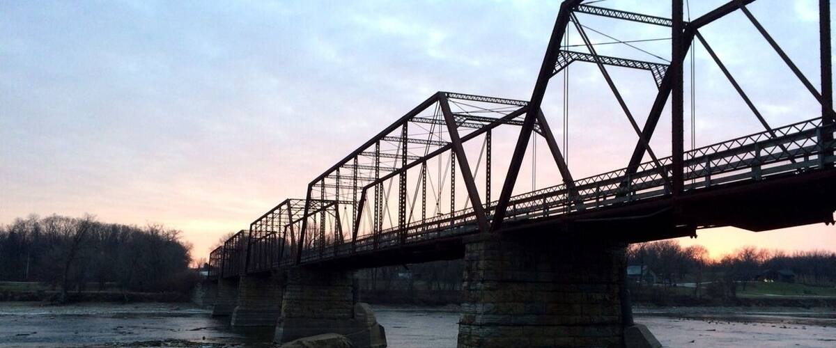 Eine Stahlträgerbrücke über einem Fluss, im Hintergrund ein Sonnenuntergang.