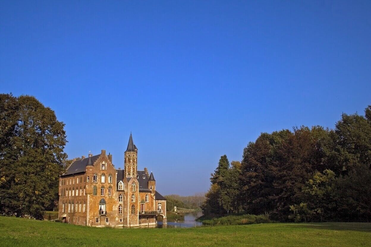A large, historic building with a prominent tower, surrounded by lush greenery and a river.