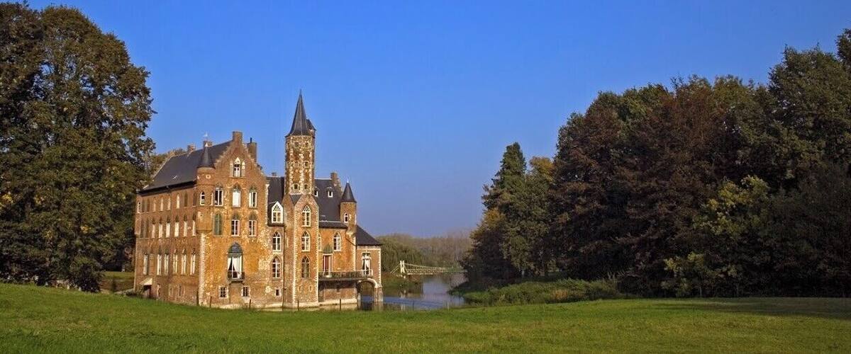 A large, historic building with a prominent tower, surrounded by lush greenery and a river.