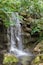 Ein Wasserfall stürzt über moosbewachsene Steine in ein kleines Becken, umgeben von dichtem Grün.
