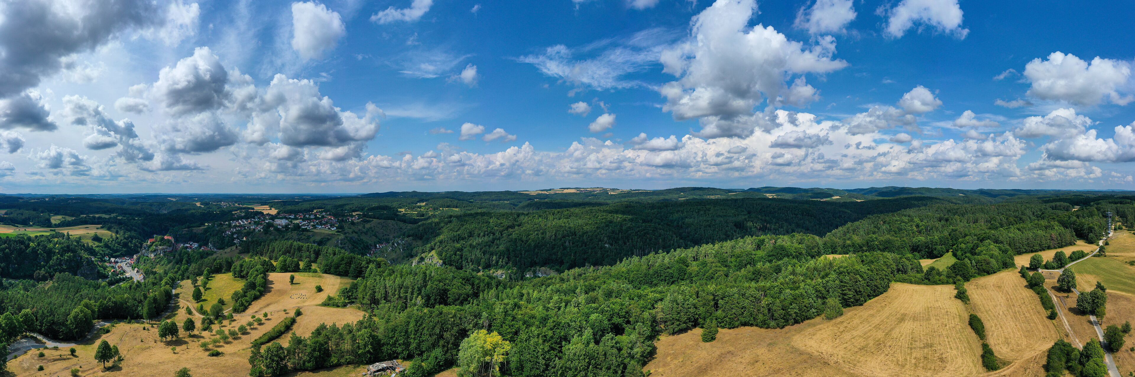 A mountainous landscape with dense forests, a small village, and a clear blue sky with scattered clouds.