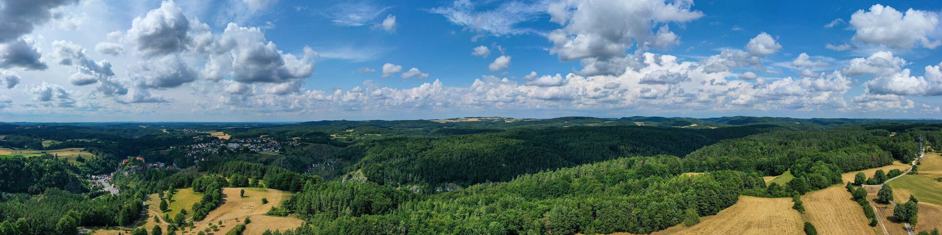 A mountainous landscape with dense forests, a small village, and a clear blue sky with scattered clouds.