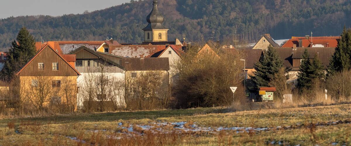 A village with traditional houses, a church with a bell tower, and a forested hill in the background.