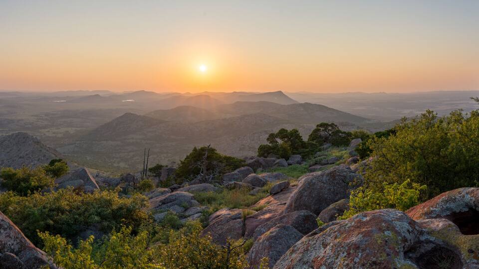 A mountainous landscape with a sunset, rocky terrain, and scattered trees.