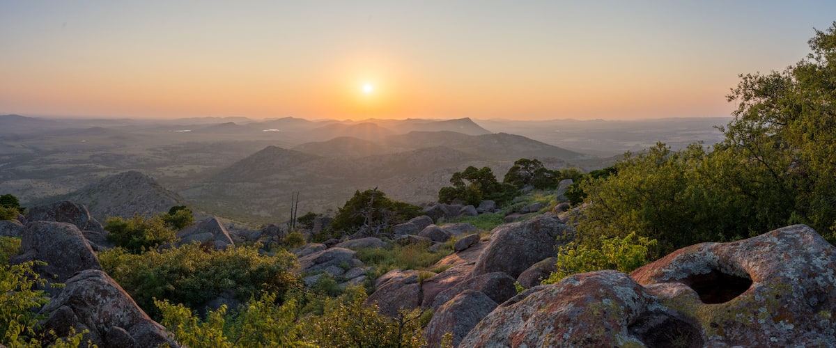 A mountainous landscape with a sunset, rocky terrain, and scattered trees.