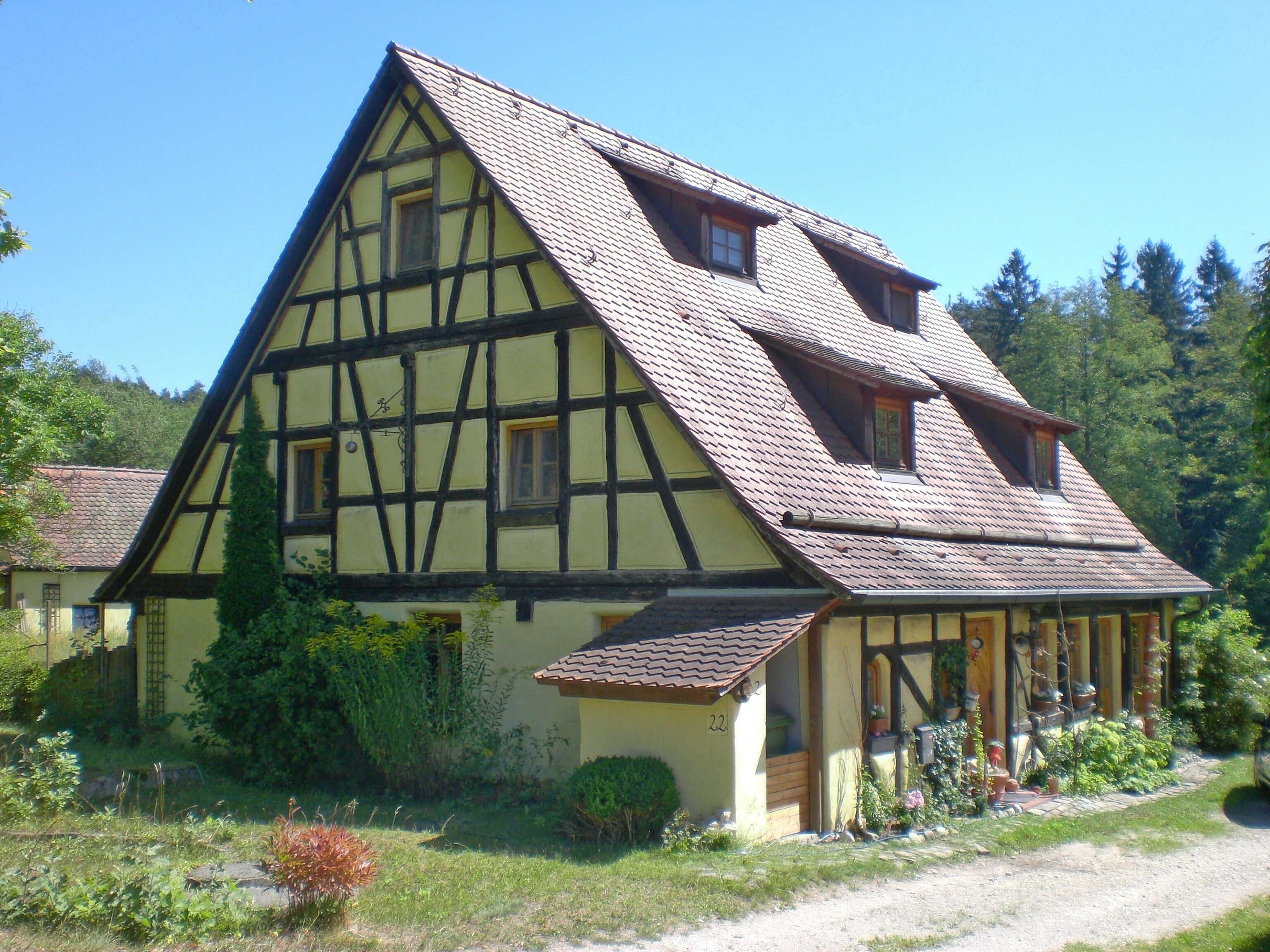 A traditional half-timbered house with a steeply pitched roof, surrounded by lush greenery and a clear blue sky.