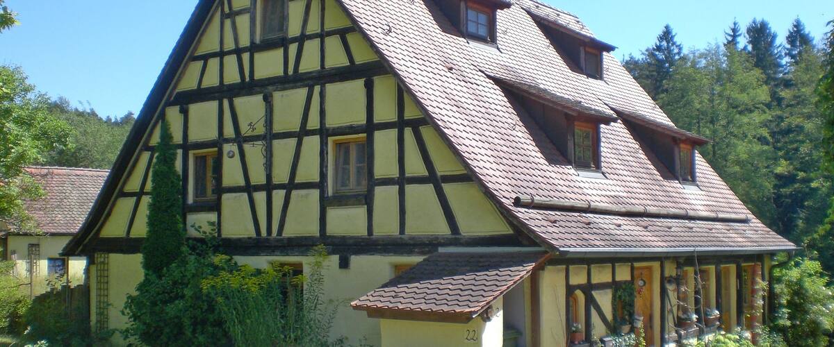 A traditional half-timbered house with a steeply pitched roof, surrounded by lush greenery and a clear blue sky.