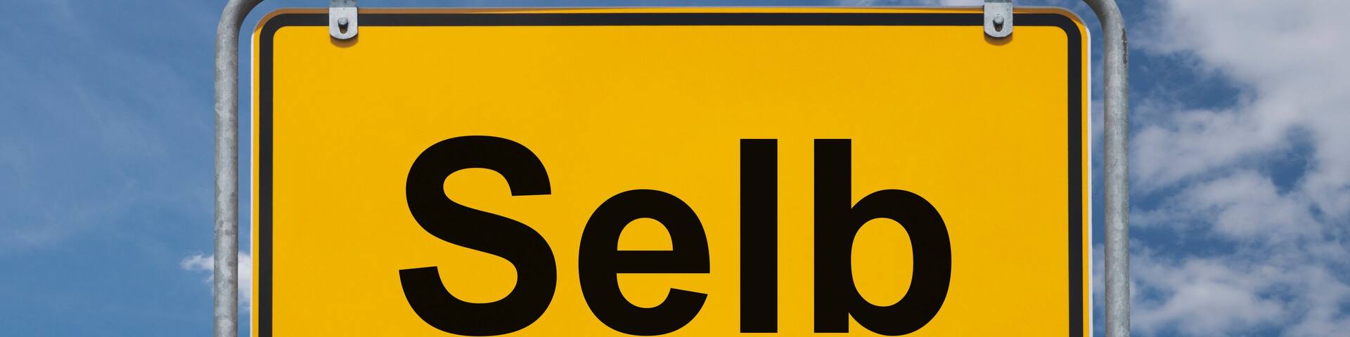 A yellow sign with the word "Selb" against a blue sky with clouds.