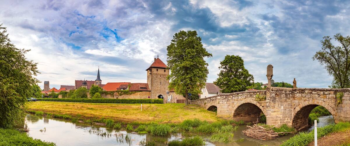 A stone bridge over a river, a church with a spire, and a tower in a town.