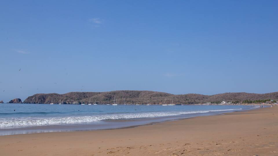 A sandy beach with calm waves, sailboats, and distant hills.