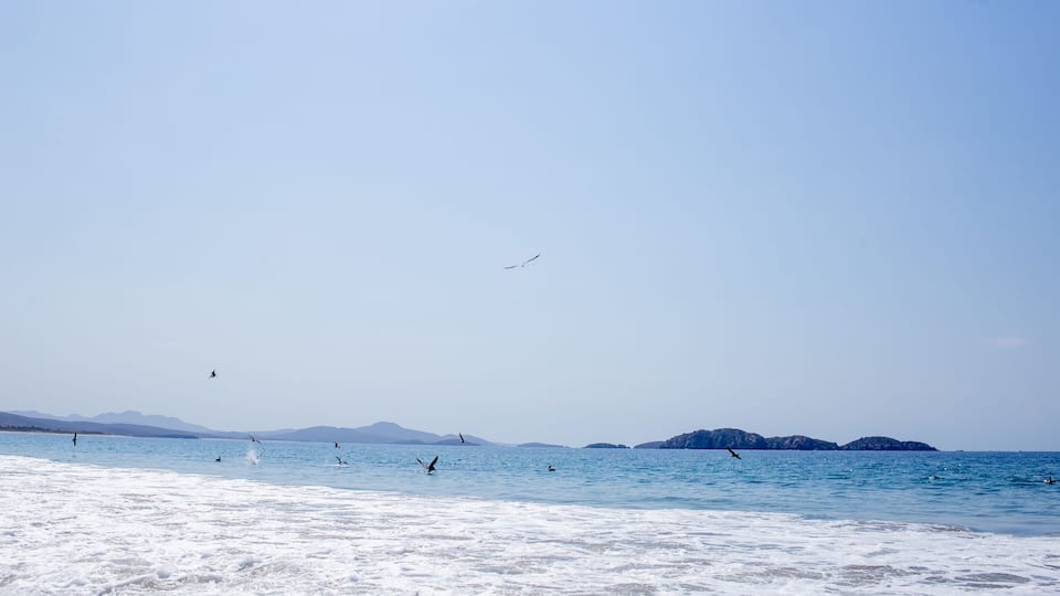 A beach with clear blue water, waves, and distant mountains.