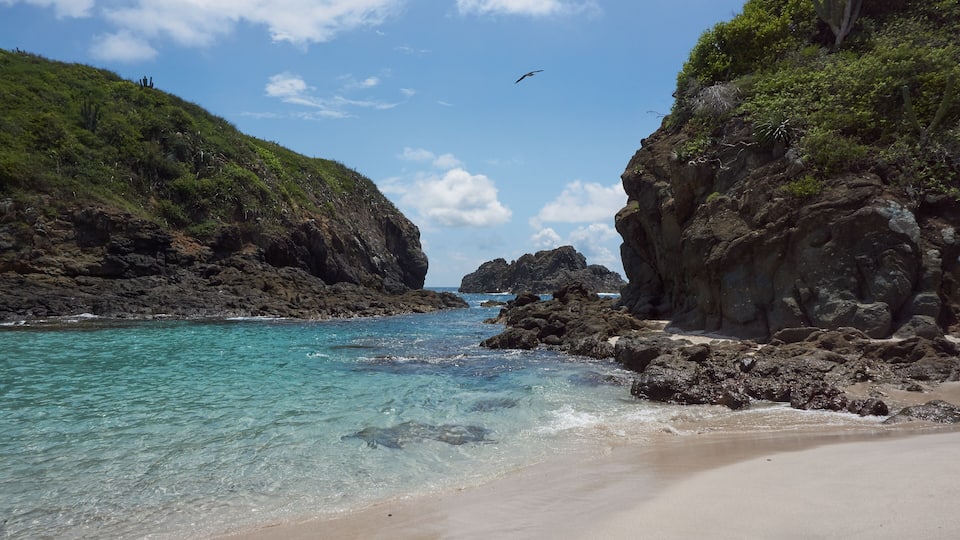 A coastal landscape with clear turquoise waters, rocky cliffs, and a sandy beach.
