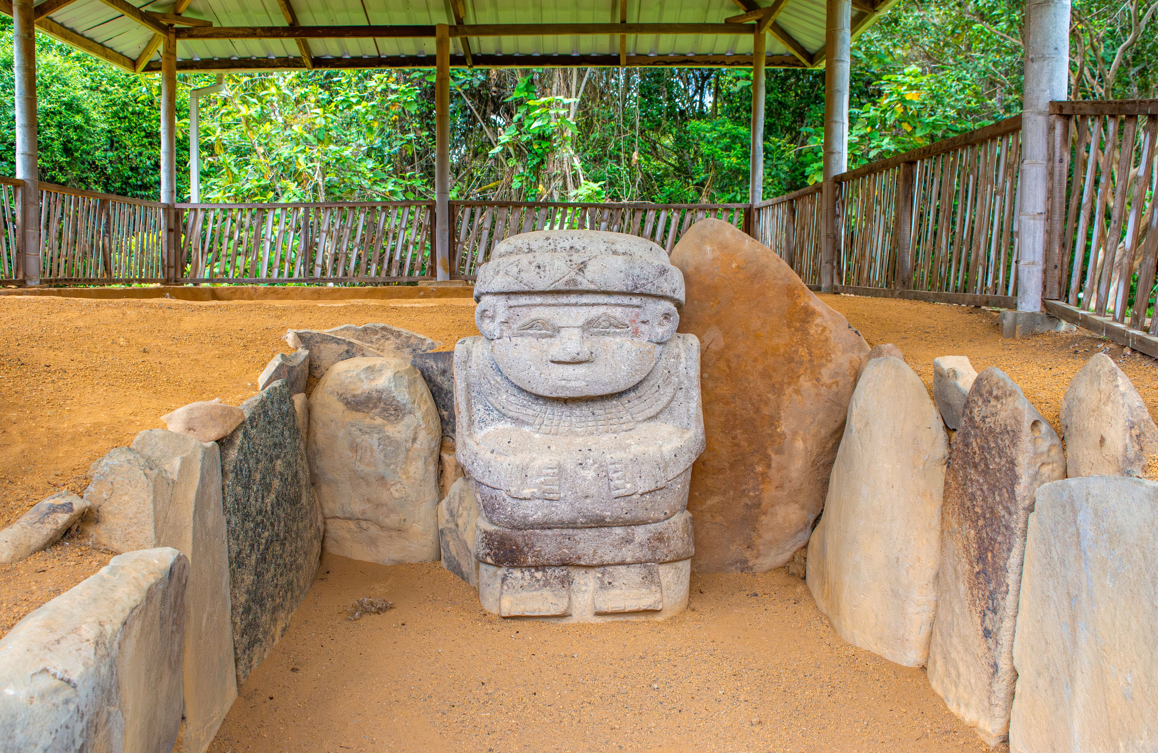 A stone statue in a sandy enclosure with a wooden fence and a covered shelter in the background.