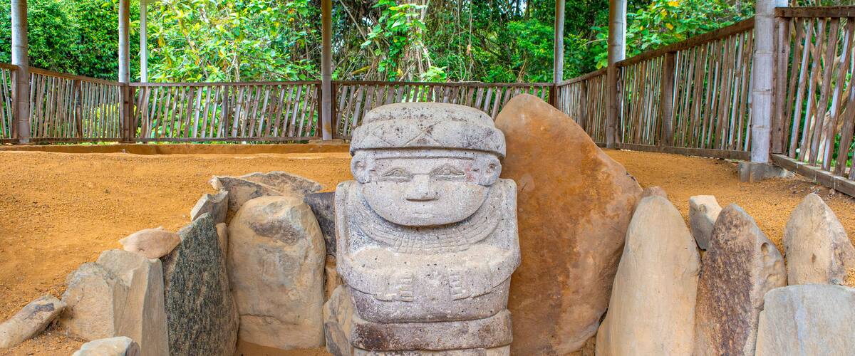 A stone statue in a sandy enclosure with a wooden fence and a covered shelter in the background.