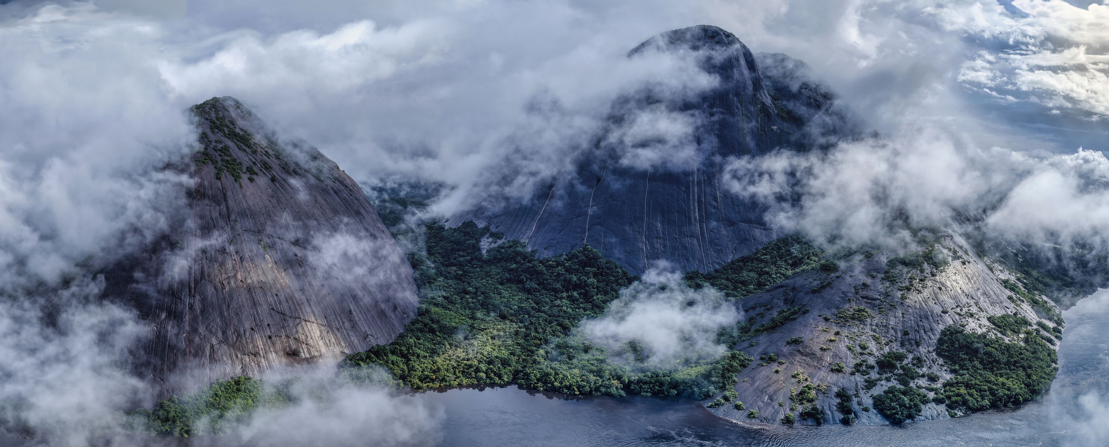 Aerial view of stark, towering rock formations piercing through a sea of swirling clouds, revealing patches of vibrant green vegetation nestled at their base, Inirida, Guainia, Colombia.