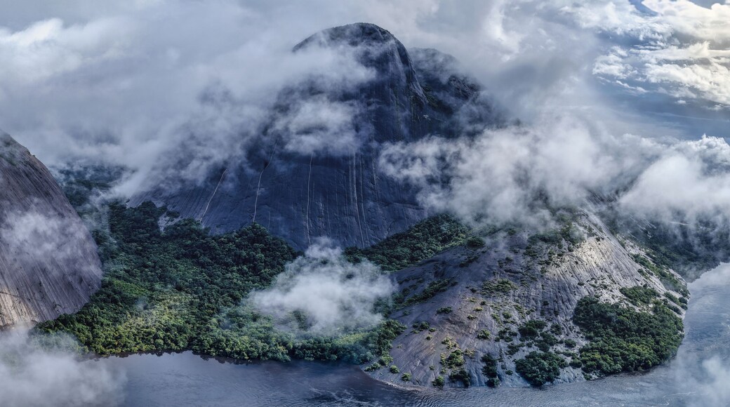 Aerial view of stark, towering rock formations piercing through a sea of swirling clouds, revealing patches of vibrant green vegetation nestled at their base, Inirida, Guainia, Colombia.