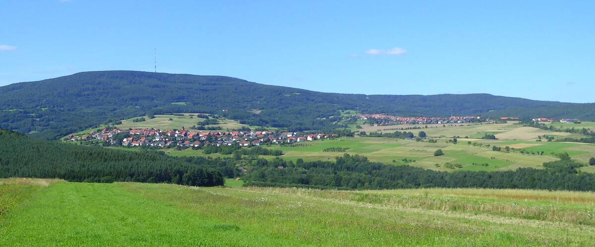 A rural landscape with rolling hills, a village with red-roofed houses, and a dense forest in the background.