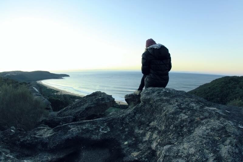 A person sitting on a rock overlooking a coastal landscape with hills in the background.