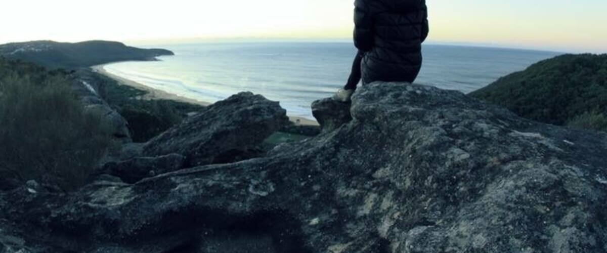 A person sitting on a rock overlooking a coastal landscape with hills in the background.