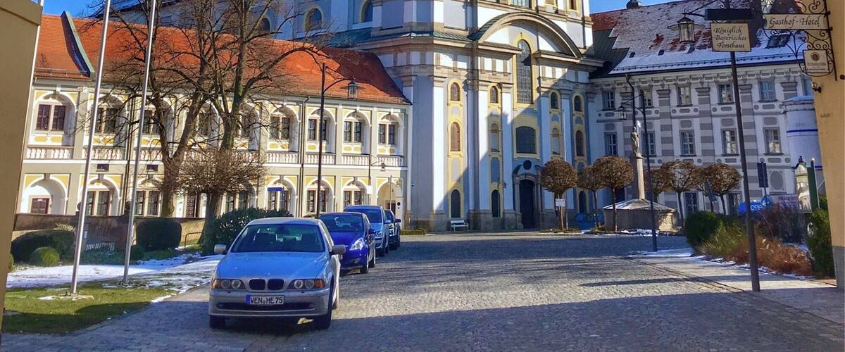 A historic church with two green domes and a clock tower, surrounded by cobblestone streets and parked cars.