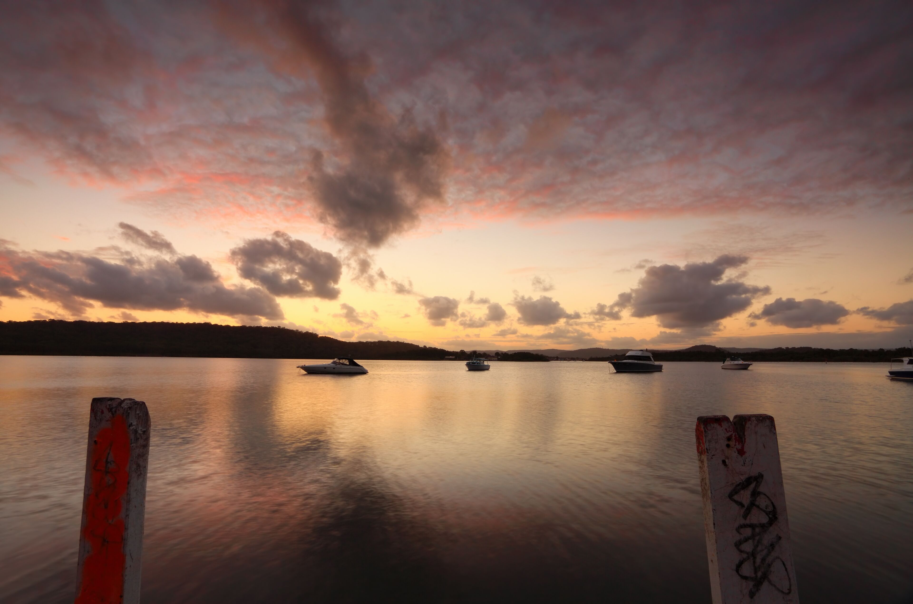 A calm lake at sunset with boats anchored in the distance and a wooden post in the foreground.
