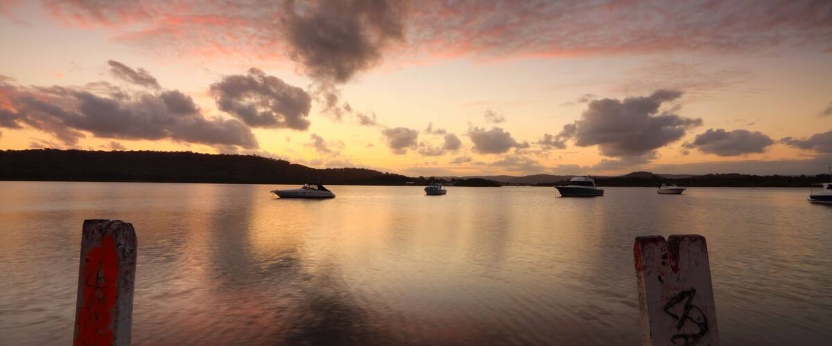 A calm lake at sunset with boats anchored in the distance and a wooden post in the foreground.