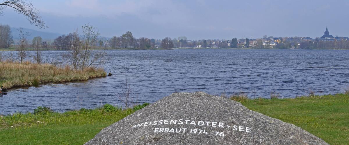 A lake with a rock inscribed with "WEISSENSTADTER ERBAUT 1974-76".