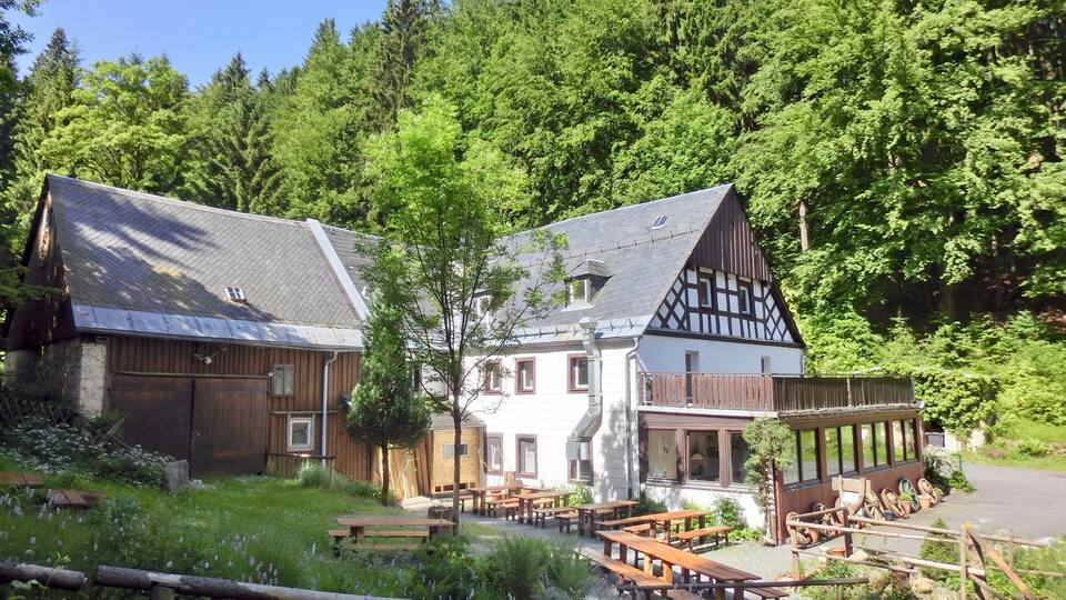 A traditional half-timbered house with a balcony, surrounded by lush greenery and a forested backdrop.