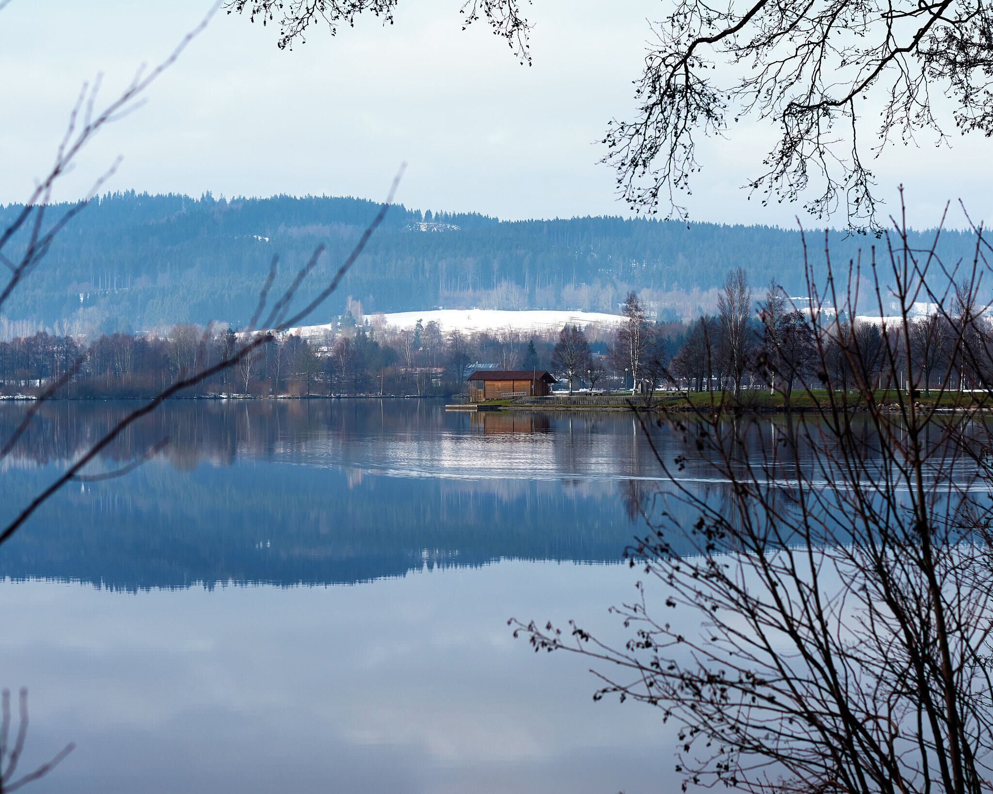 A calm lake with a wooden cabin on the shore, surrounded by trees and a snow-covered hill in the background.