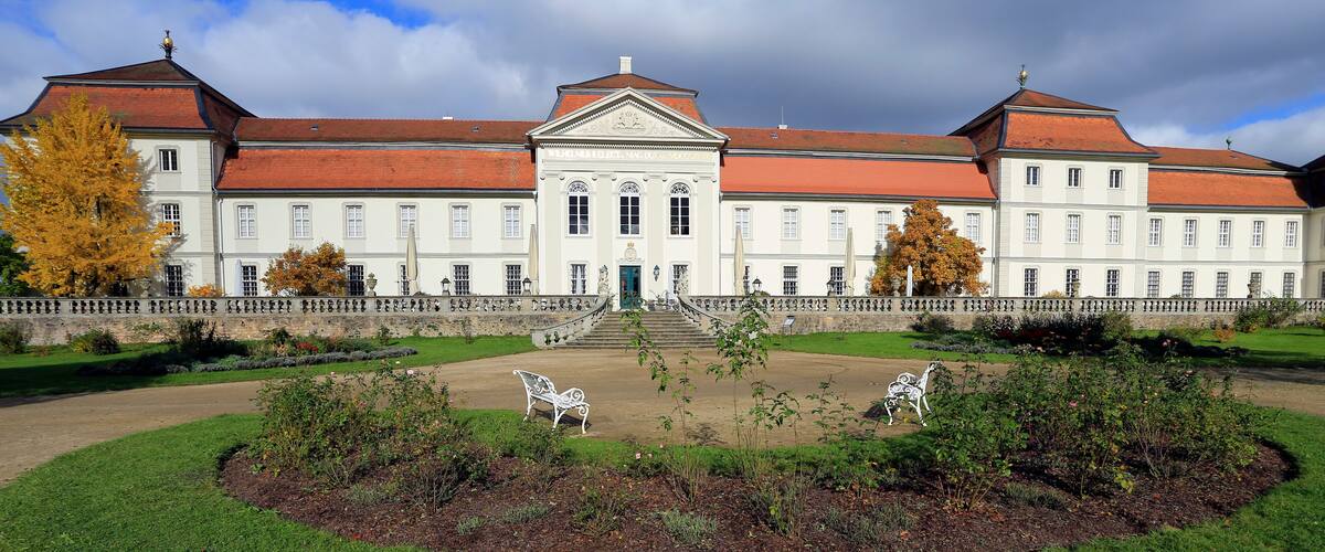 A large, historic building with a red roof and white walls, surrounded by a well-maintained garden with a central pathway and two white statues.
