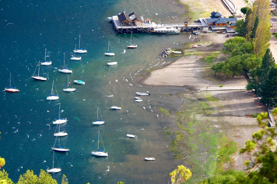 Zona costera con numerosos veleros anclados, un muelle con edificios y una playa de arena con árboles.