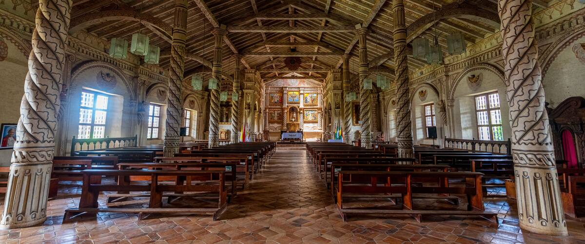 Interior de una iglesia con bancales de madera, piso de baldosas y un techo alto con diseños intrincados.