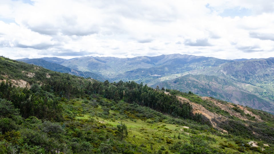 A mountainous landscape with dense green vegetation and distant mountain ranges under a cloudy sky.