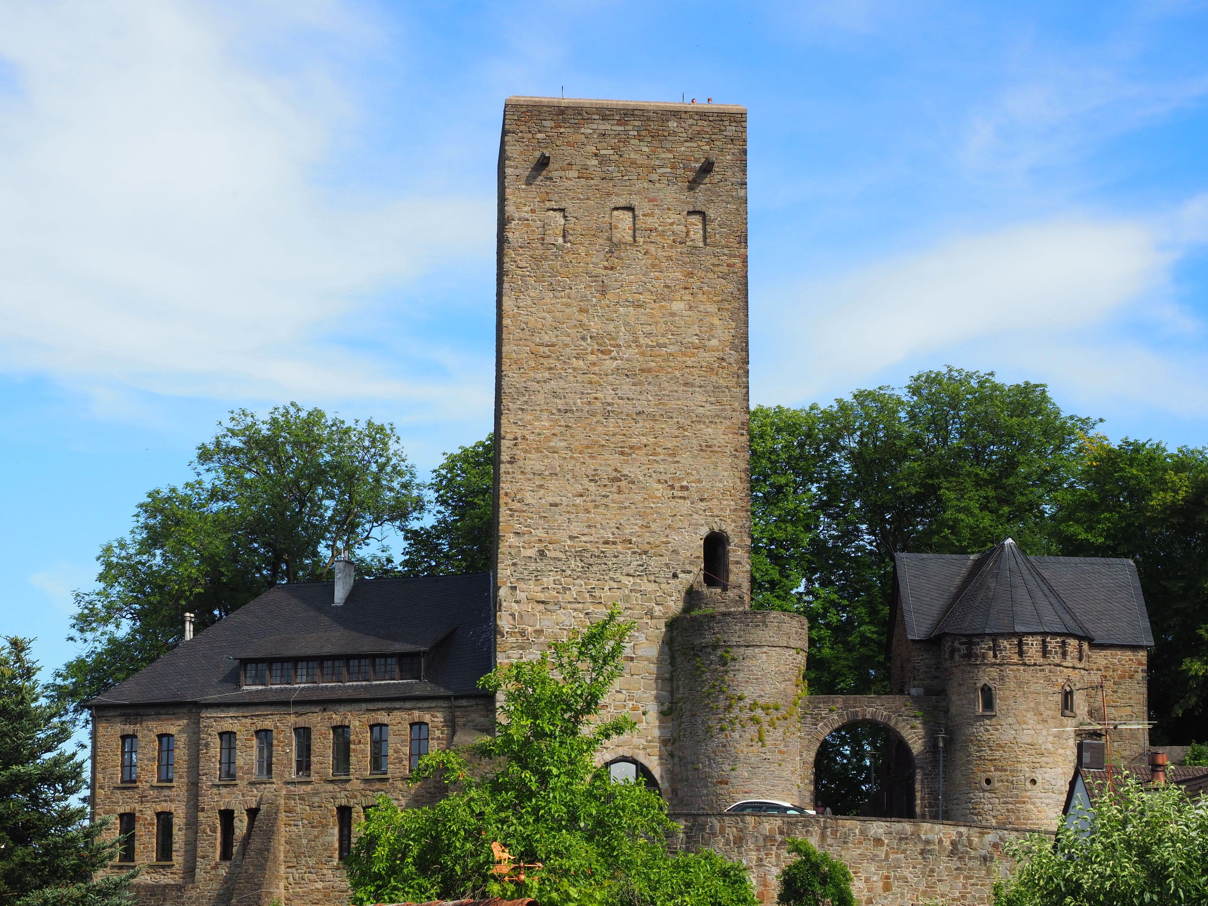 A historic stone tower with arched windows, a large building with multiple windows, and lush green trees in the background.